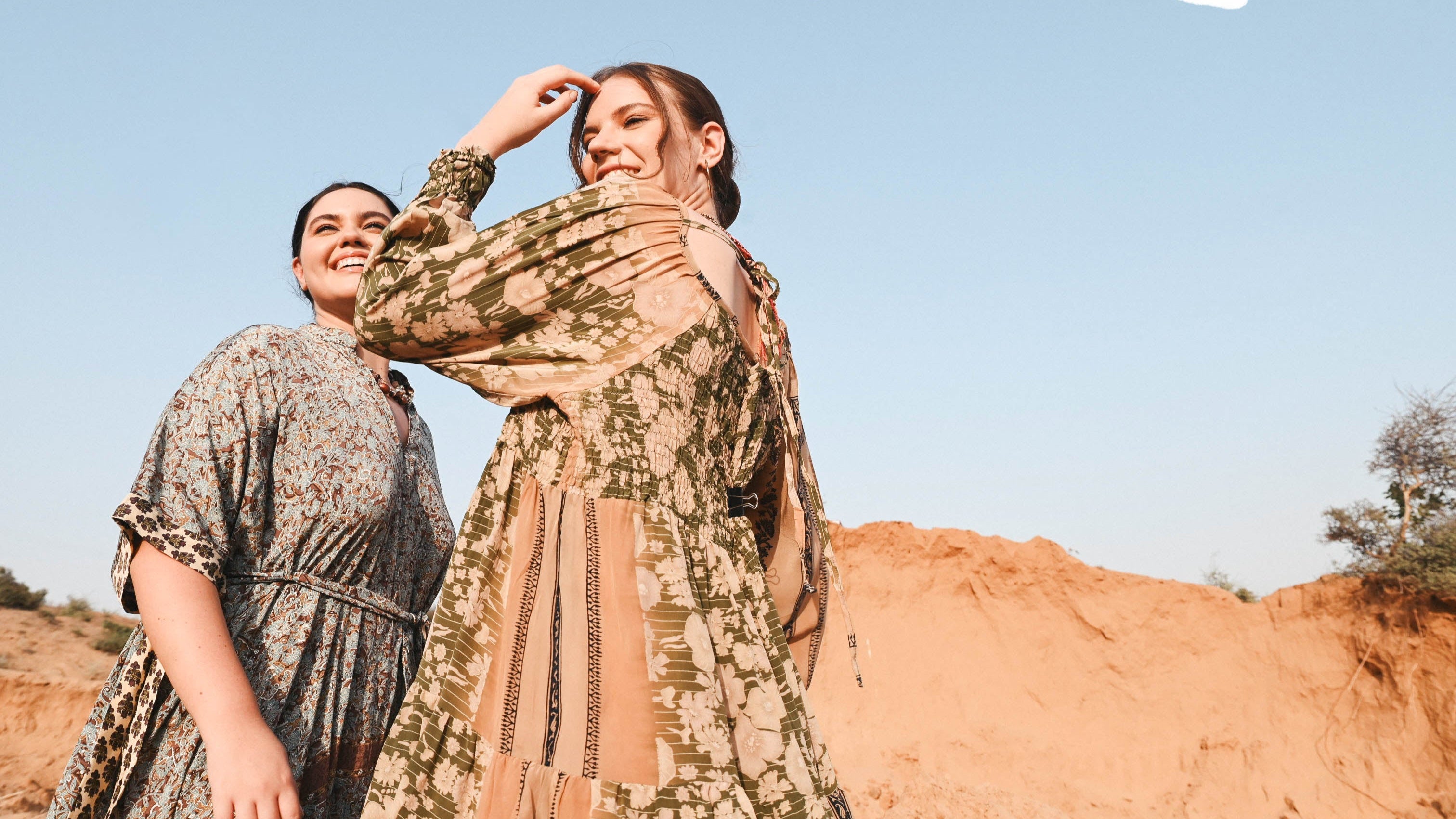 Two people in patterned outfits standing in a desert landscape with mountains in the background.